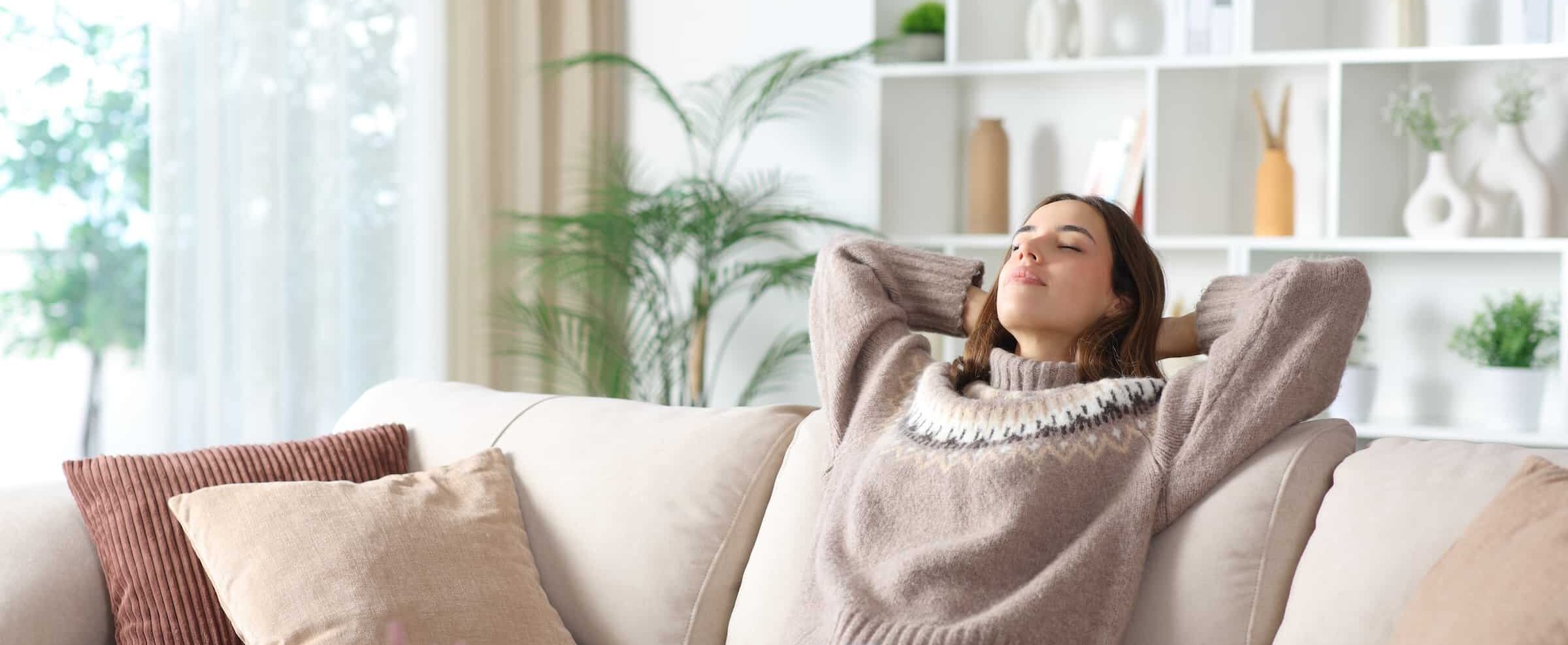 Woman relaxed with her arms behind her head on the living room couch. She has her eyes closed and is breathing peacefully.